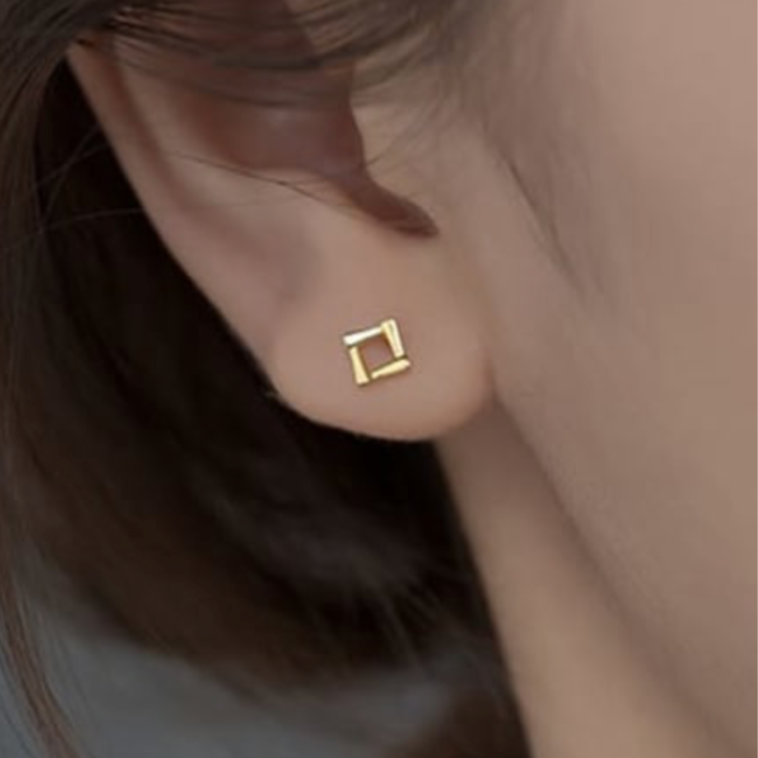 Gold square earring worn on an ear with a neutral background