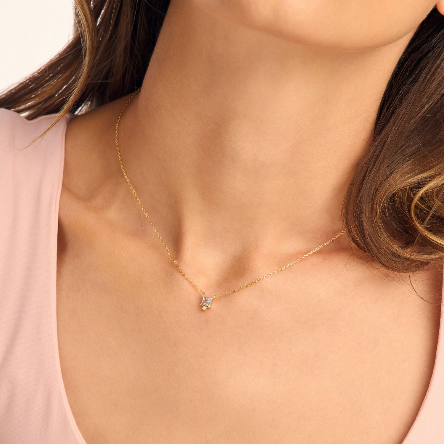 Model wearing a blue CZ drop necklace, featuring a small white stone and a vibrant blue cubic zirconia on a gold chain for an elegant, timeless look