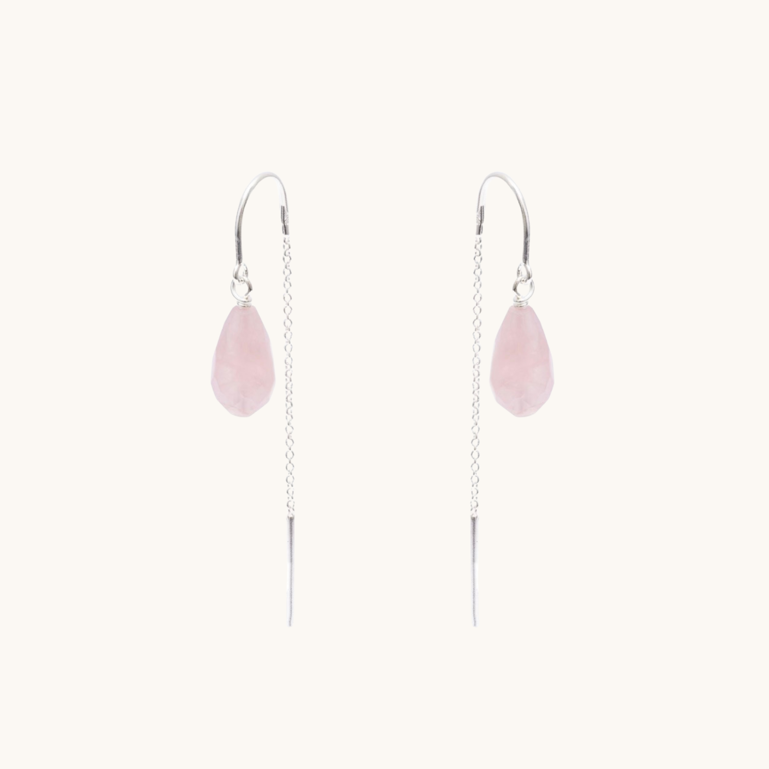 A pair of long dangle earrings featuring a light pink teardrop-shaped stone suspended from a silver wire chain with a silver hook back, set against a solid off-white background.