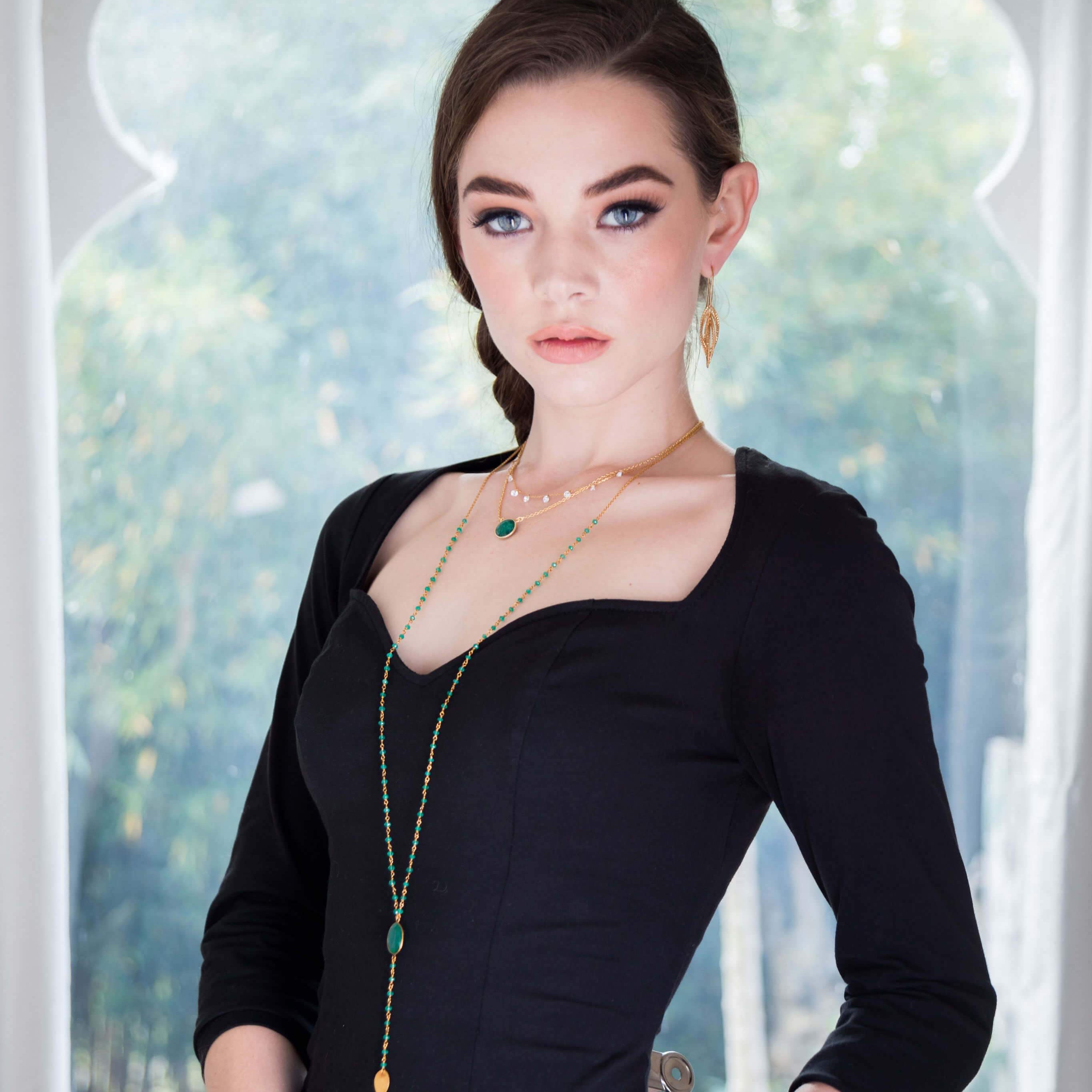 Model wears gold plated necklace with green semi-precious stones layered with other pieces